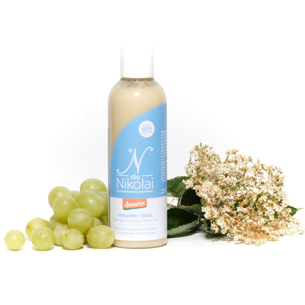 dieNikolai Holunder Duschgel und Shampoo in 100% recycelter Flasche mit blauem N-Etikett, daneben grüne Trauben und frische Holunderblüten – Demeter Bio-Naturkosmetik aus der Wachau