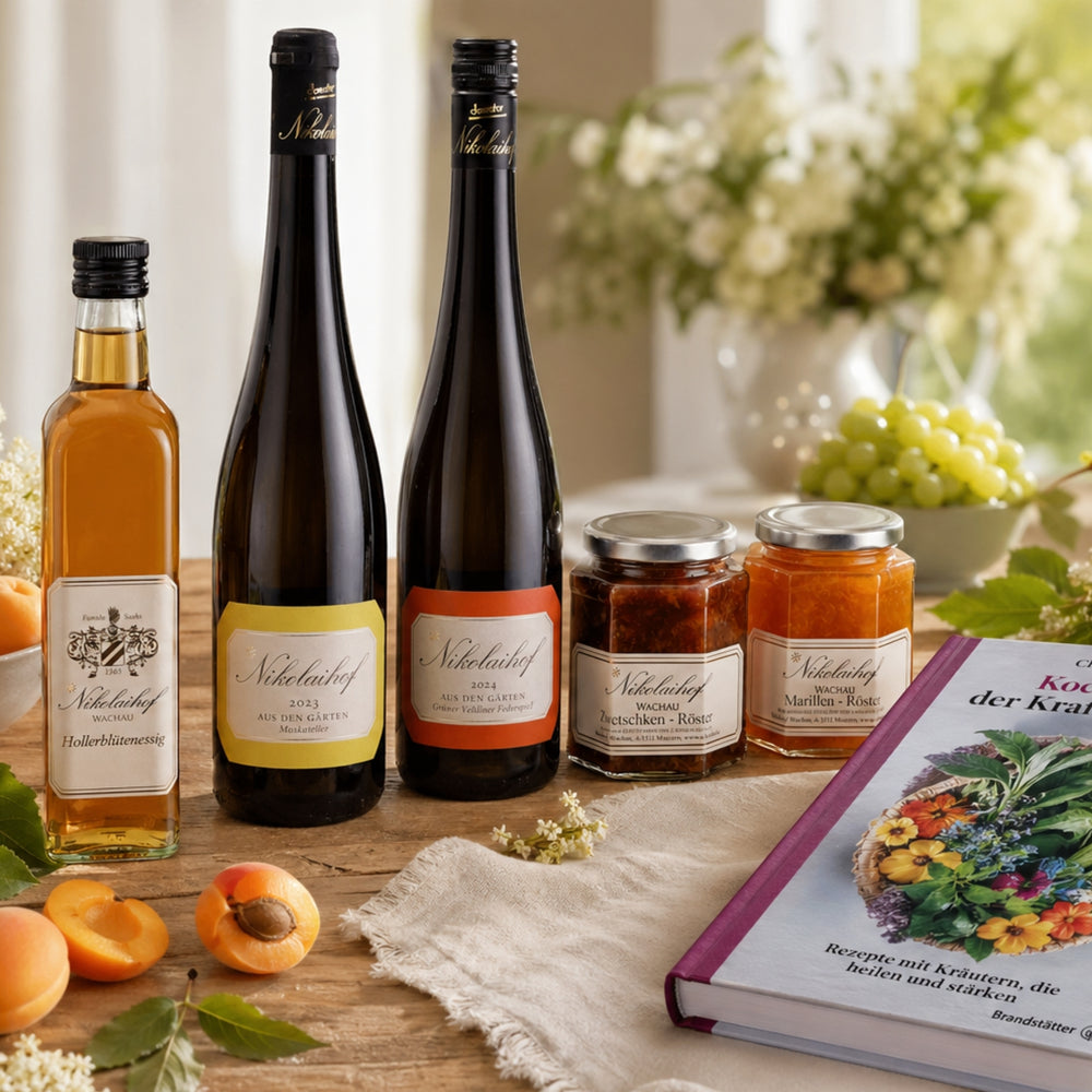 Tischarrangement mit Nikolaihof Wachau Hollerblütensirup, Weinflaschen, Bio-Marmeladen und Kochbuch der Kräuter neben Marillen – Mitbringsel aus dem Sortiment von dieNikolai aus Österreich