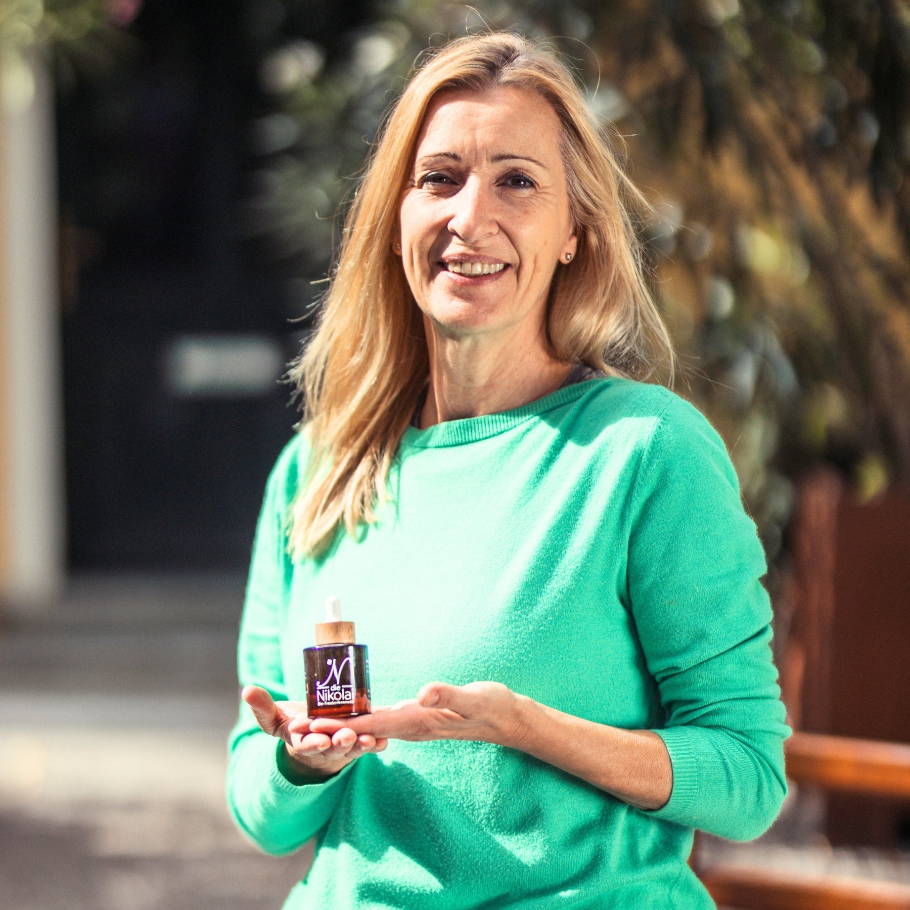 Blonde Frau im grünen Pullover präsentiert lächelnd ein dunkles Fläschchen Traubenkern Intensiv Serum – dieNikolai Demeter Bio-Traubenkosmetik aus der Wachau in Österreich