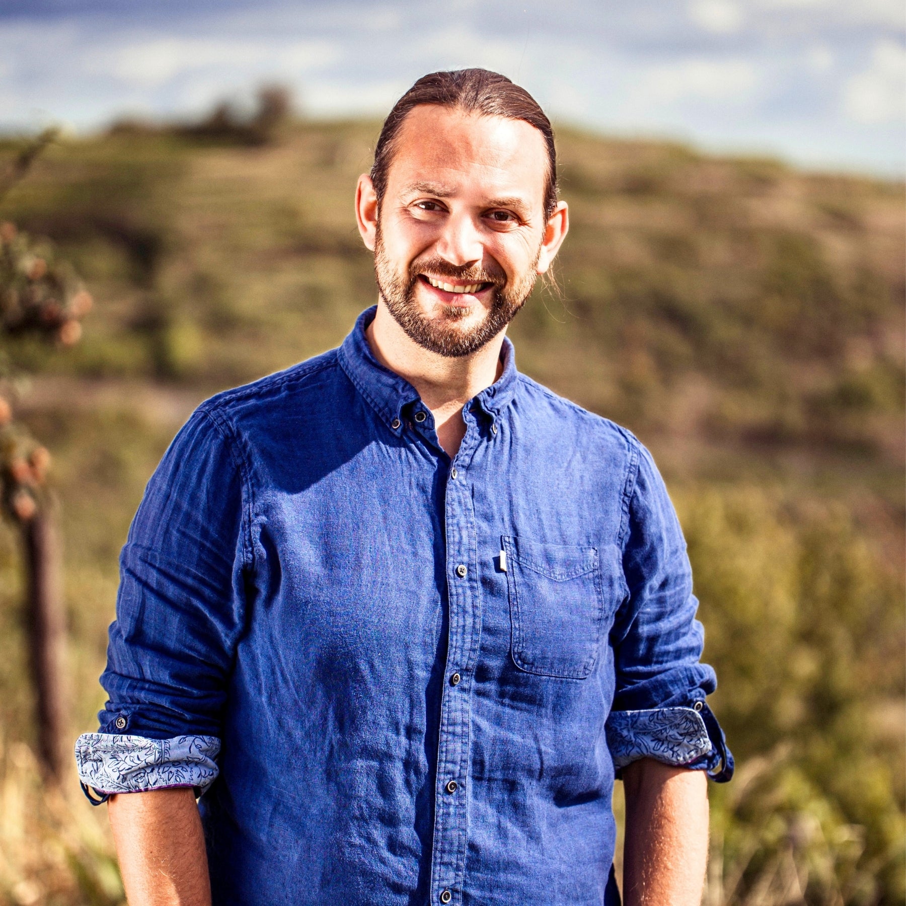 Lächelnder Mann mit Bart im blauen Leinenhemd vor Weingärten der Wachau – Portrait aus dem Team von dieNikolai Bio-Traubenkosmetik aus Österreich