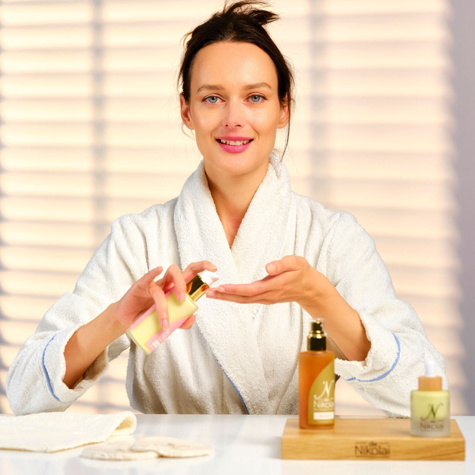 Frau im Bademantel verteilt Pflegeprodukt auf der Handfläche, vor ihr stehen dieNikolai Reinigungsmilch und Fluid – Wellness mit Demeter Bio-Traubenkosmetik aus der Wachau