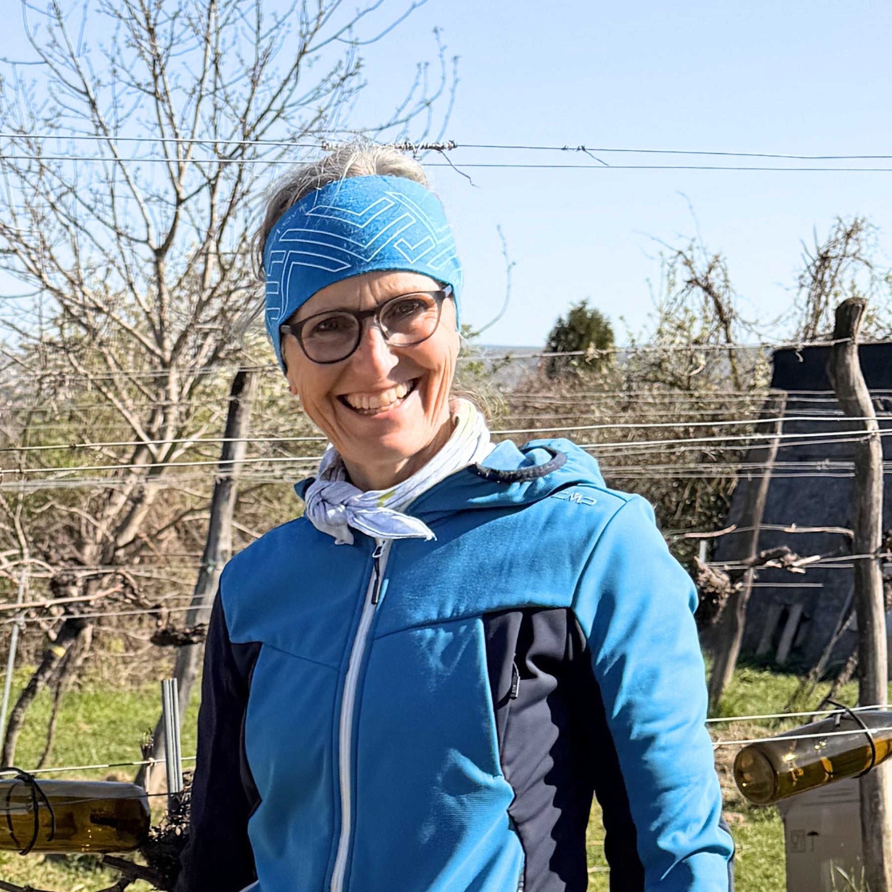 Lächelnde Winzerin mit Brille und blauem Stirnband im Frühlings-Weingarten der Wachau – authentische Bio-Traubenkosmetik vom Nikolaihof bei dieNikolai