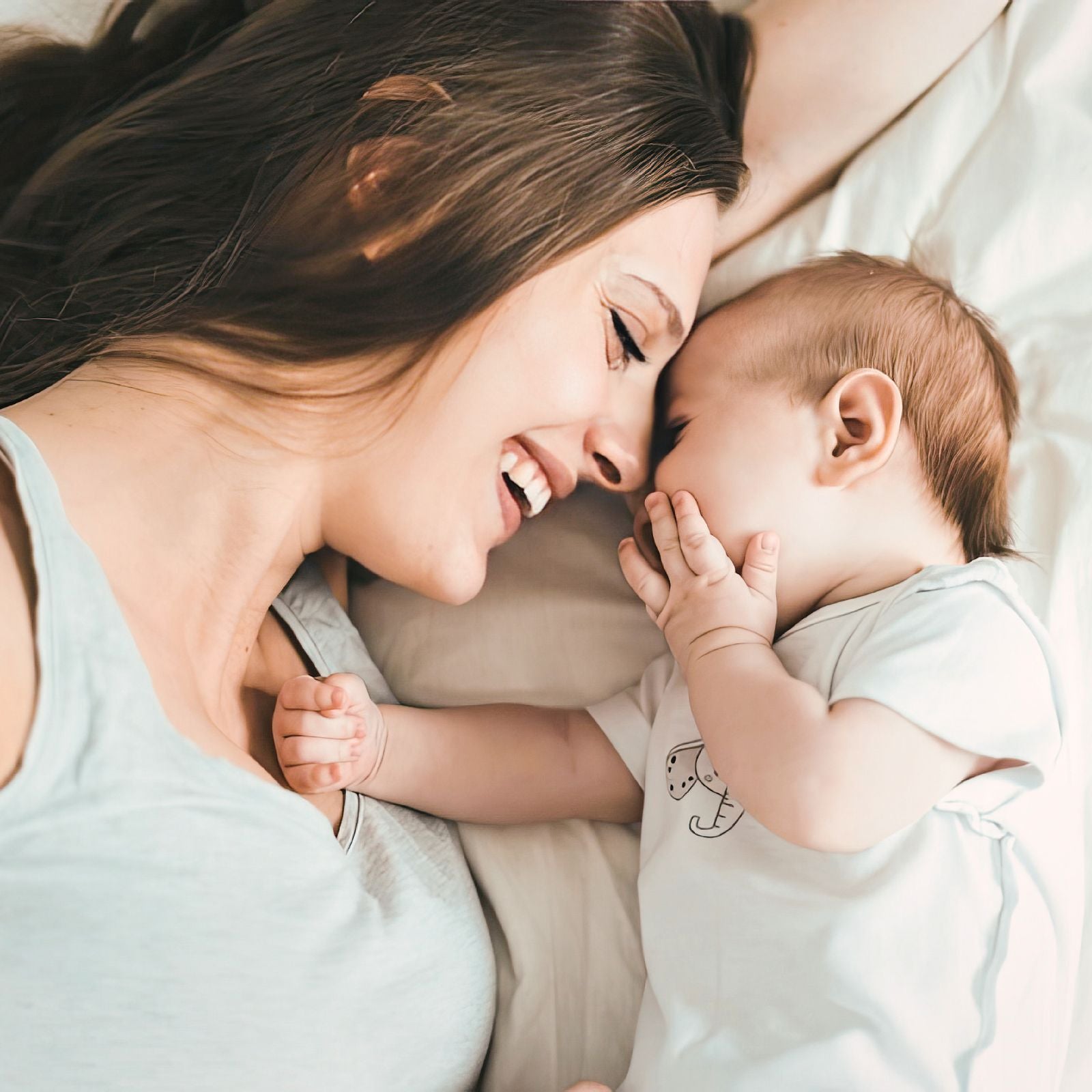 Junge Mutter und Baby liegen Stirn an Stirn im weißen Bett und lachen – innige Momente passend zur dieNikolai Baby & Kids Bio-Traubenkosmetik aus der Wachau