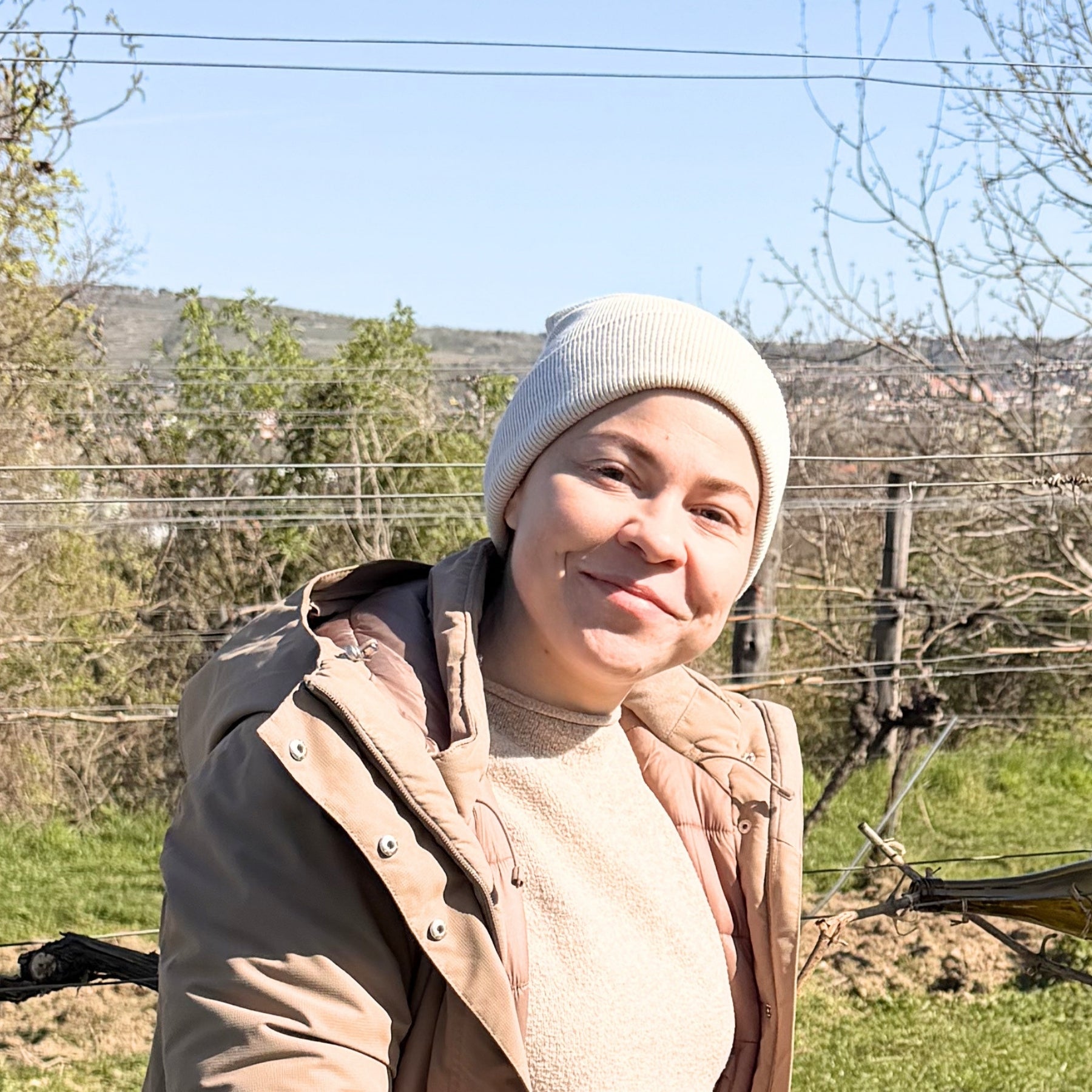 Lächelnde Frau mit hellem Beanie und beiger Steppjacke im Weingarten der Wachau – persönliches Porträt hinter dieNikolai Demeter Bio-Traubenkosmetik aus Österreich