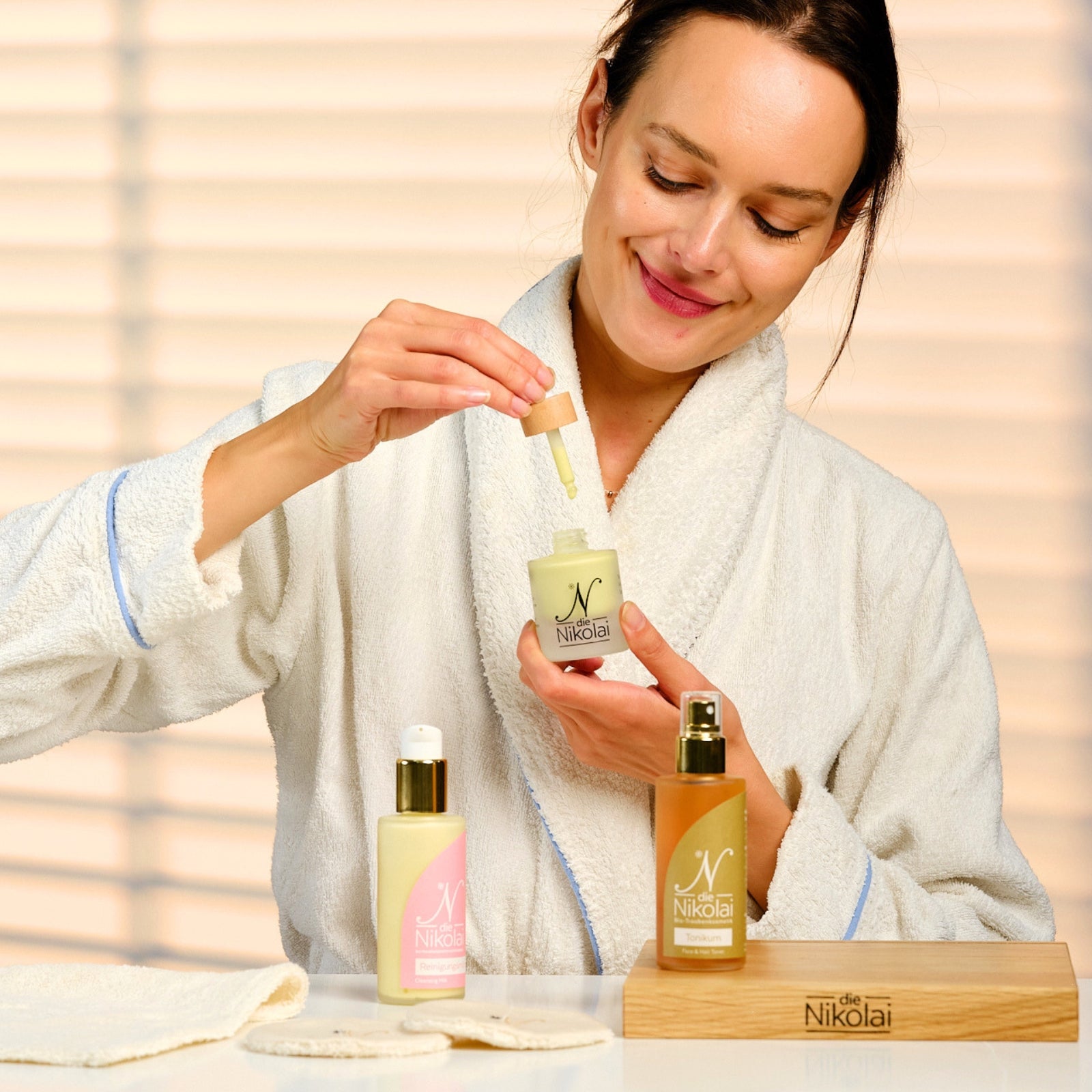Frau im Bademantel träufelt Traubenkern Intensiv Serum aus Pipette, davor dieNikolai Reinigungsmilch und Tonikum – Demeter Bio-Traubenkosmetik aus der Wachau