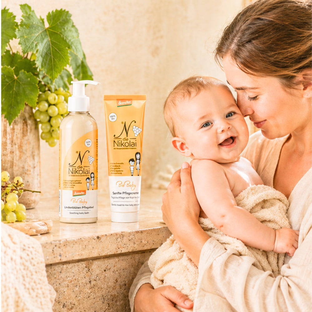 Mutter hält lächelndes Baby neben dieNikolai BioBaby Unterstützen-Pflegeöl und Sanfter Pflegecreme – Demeter Bio-Traubenkosmetik für Babypflege aus der Wachau