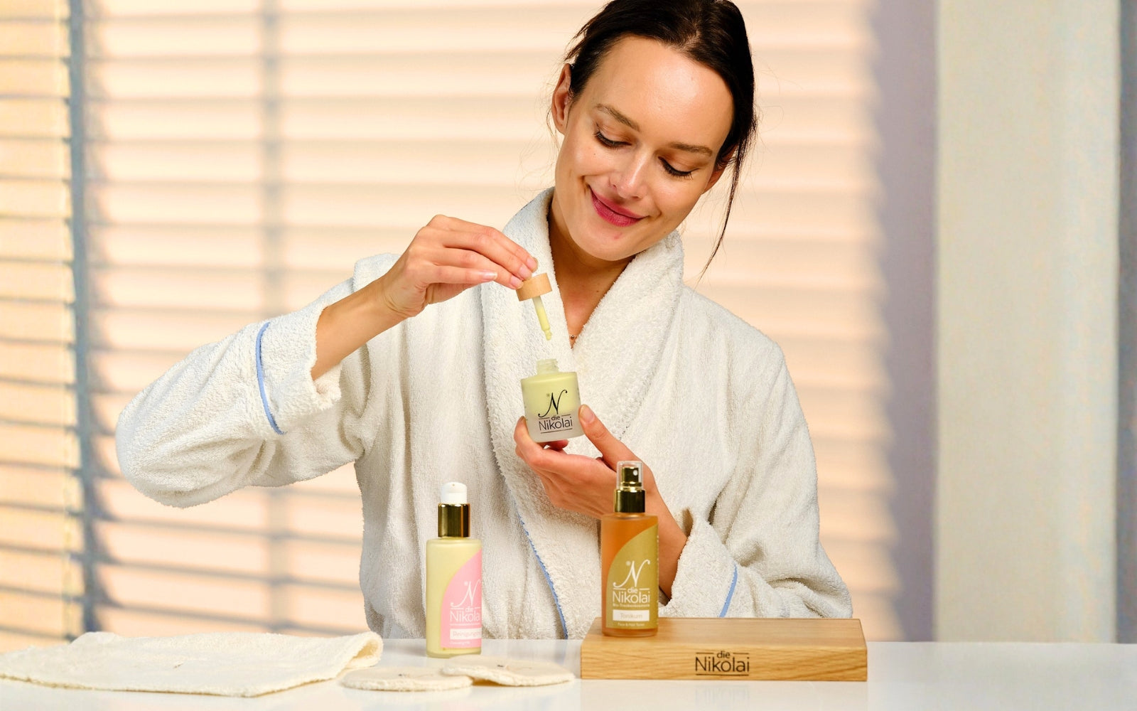 Frau im weißen Bademantel träufelt dieNikolai Intensiv-Serum auf die Hand, daneben Lieblingsfluid und Gesichts-Tonikum – Demeter Bio-Traubenkosmetik aus der Wachau