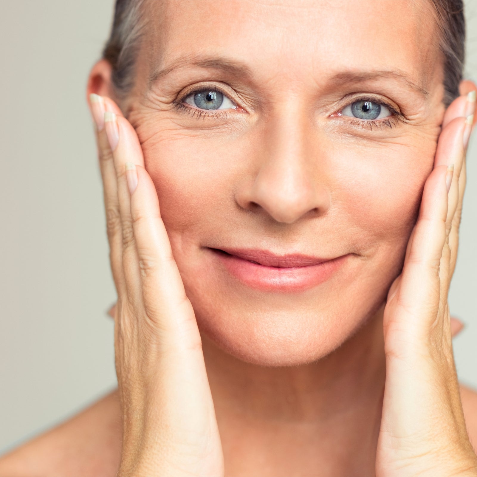 Reife Frau mit strahlend gepflegter Haut umfasst lächelnd ihre Wangen – Anti-Aging Wirkung der dieNikolai Demeter Bio-Traubenkosmetik aus der Wachau