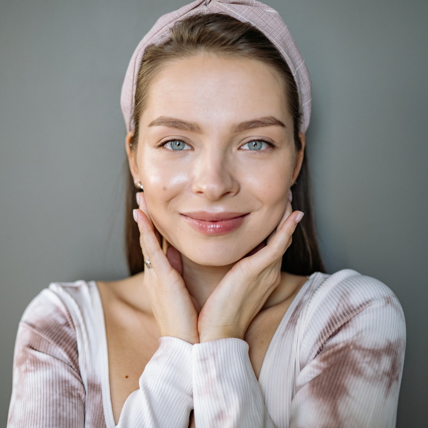 Junge Frau mit rosa Stirnband und makelloser Haut umrahmt ihr Gesicht mit den Händen – natürliche Reinheit mit dieNikolai Demeter Bio-Traubenkosmetik aus der Wachau
