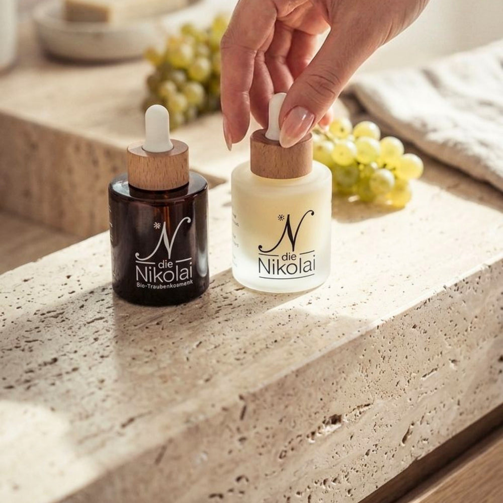 Hand greift nach zwei dieNikolai Serum-Flakons in Braun- und Milchglas auf Travertinblock mit Trauben – Demeter Bio-Traubenkosmetik aus der Wachau in Österreich
