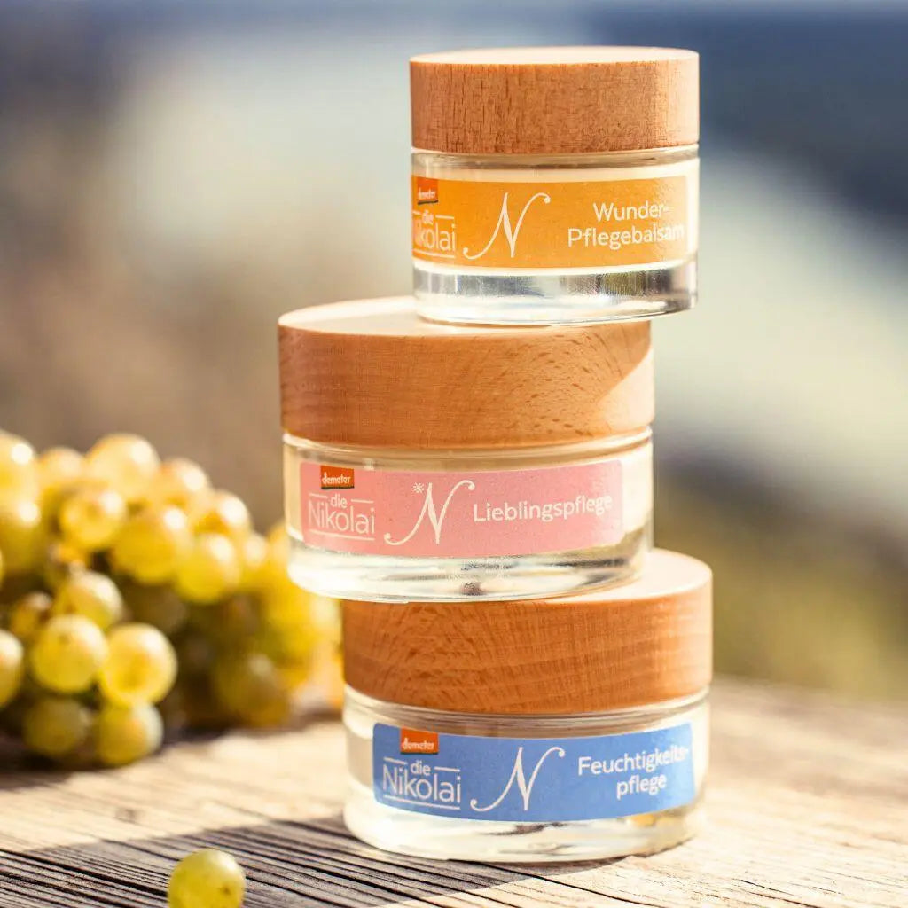 Drei gestapelte dieNikolai Tiegel Wunder-Pflegebalsam, Lieblingspflege und Feuchtigkeitspflege mit Holzdeckel vor unscharfen Weintrauben – Demeter Bio-Traubenkosmetik aus der Wachau