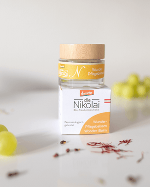dieNikolai Wunder-Pflegebalsam 30ml im Glastiegel mit Holzdeckel und Verpackung neben Safran-Fäden und Trauben – Demeter Bio-Spezialpflege aus der Wachau